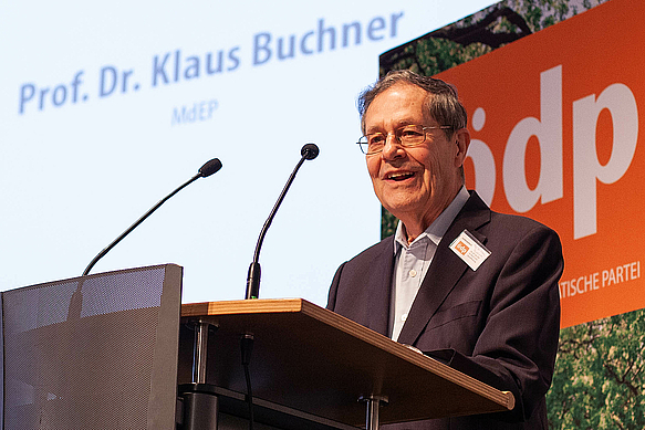 Dr. Klaus Buchner steht an einem, Rednerpult. Im Hintergrund ist eine große ÖDP Fahne aufgehängt und sein Name ist mit einem Beamer an die Wand geworfen.