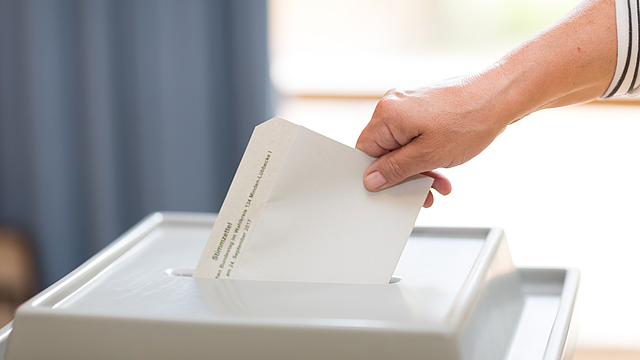 Eine Frauenhand steckt einen Briefumschlag in eine Wahlurne.