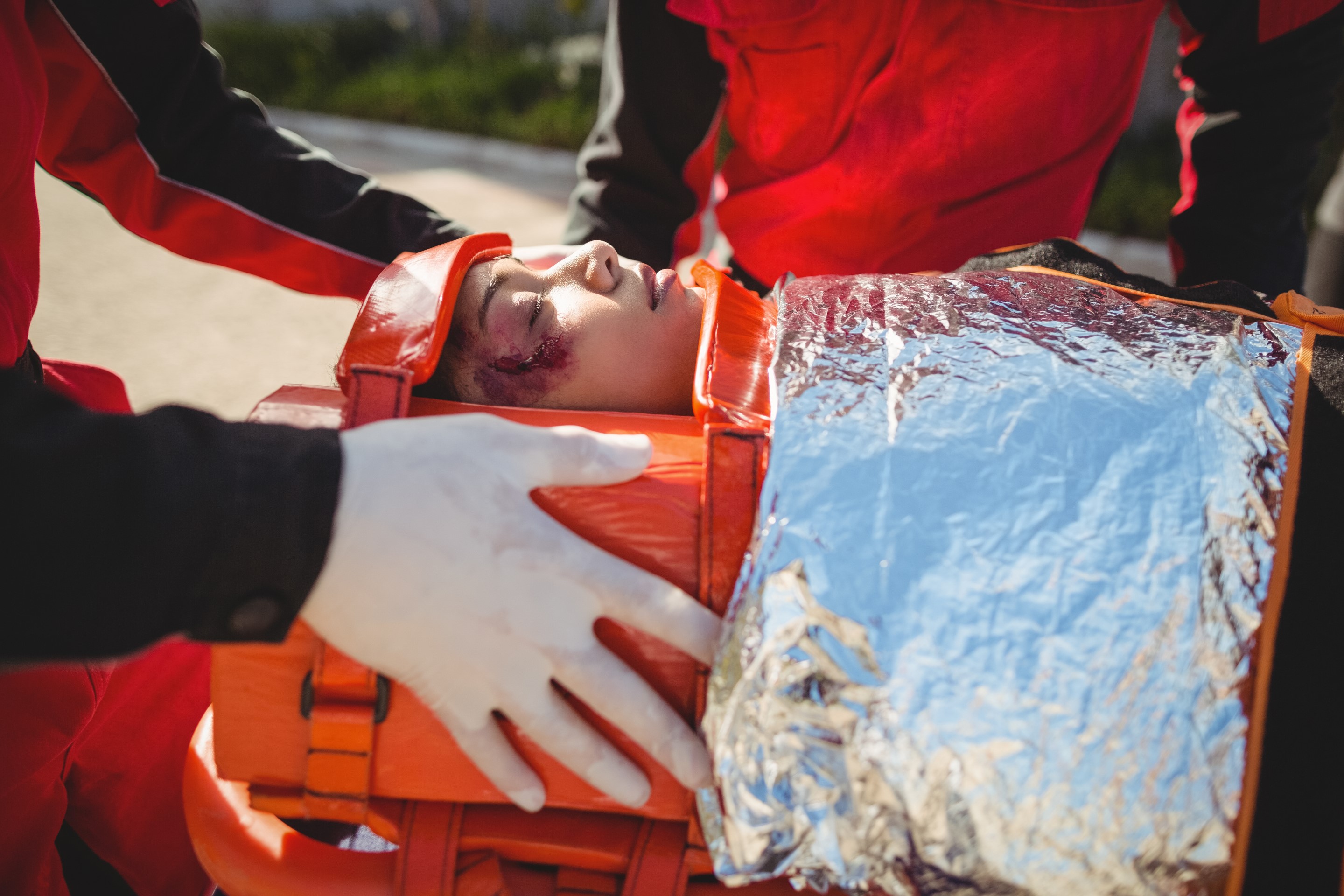 Eine junge Frau liegt in einer speziellen Fixierung auf dem Rücken. Sie hat eine blutige Wunde im Gesicht, da sie offenbar einen Unfall hatte. Zwei Personen in Uniformen von Rettungskräften kümmern sich um die Frau.
